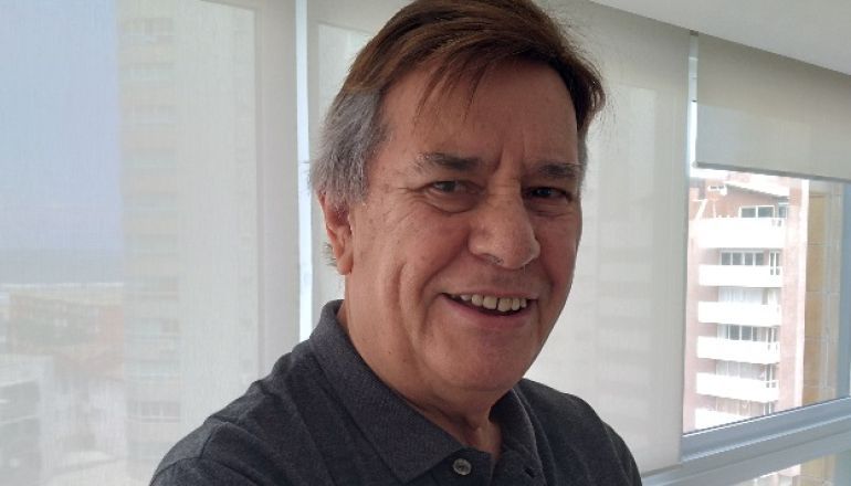 Um homem de camisa cinza está sorrindo em frente a uma janela.