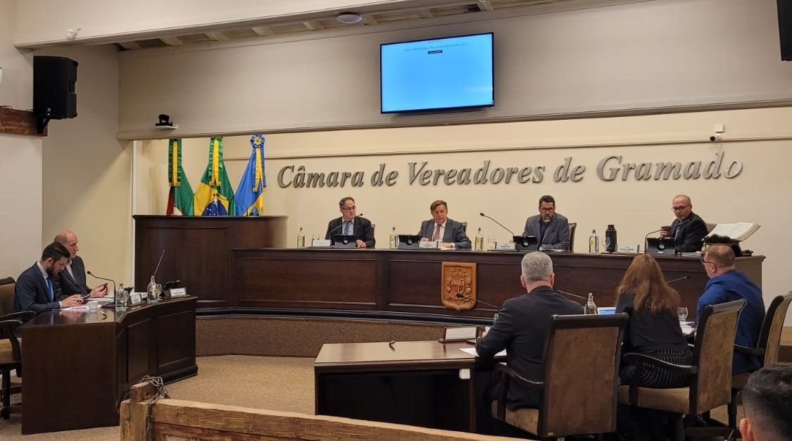 Um grupo de pessoas está sentado em mesas em uma sala com uma placa que diz camara de vereadores de gramado.