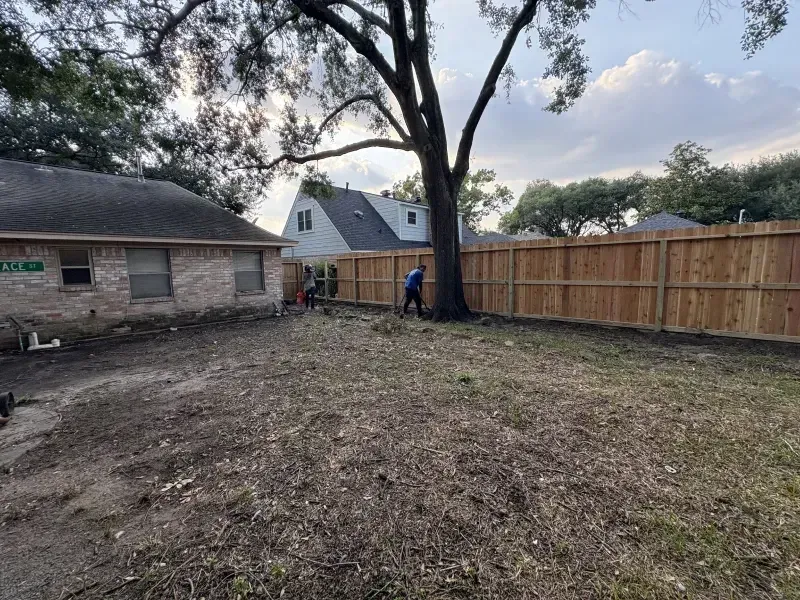 Backyard with new wooden fence, large tree, and person working. A brick house is on the left.
