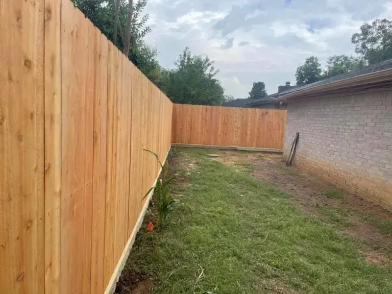 A wooden fence surrounds a grassy backyard, adjacent to a brick house. Overcast sky.