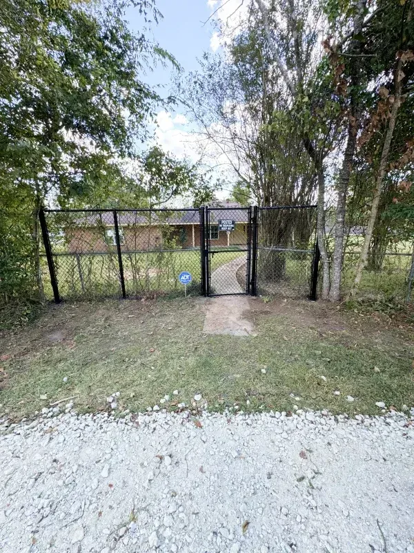 Black fence with gate leading to a house. Concrete path in front of the gate, gravel ground.