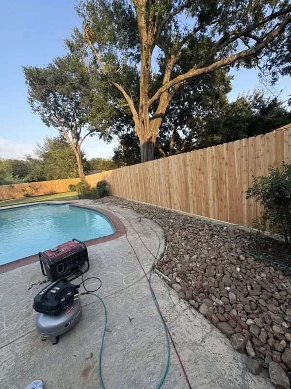 Poolside view with a wooden fence, large tree, and landscaping. A generator and air compressor are visible.