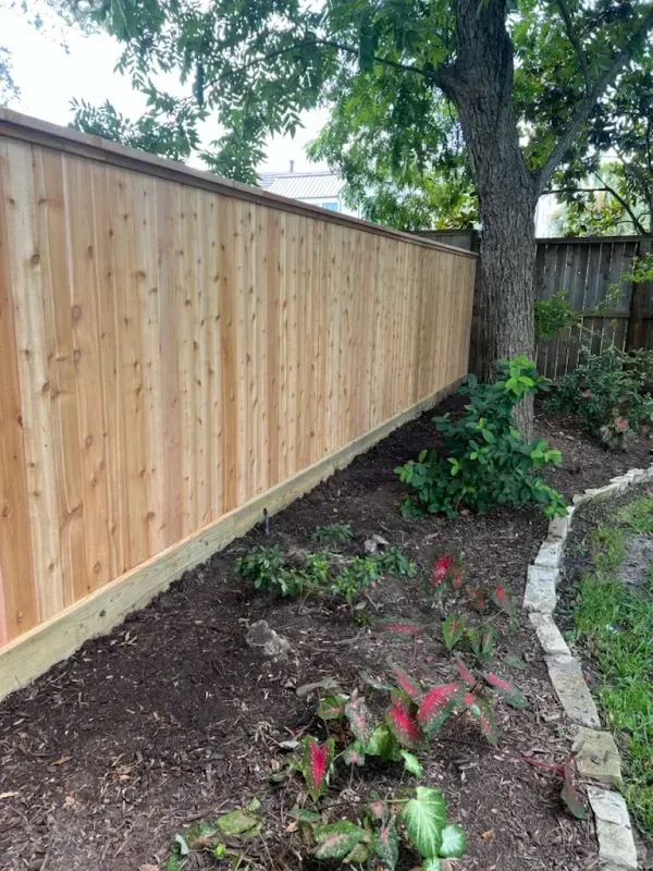 Wooden fence along a garden bed with plants, mulch, and a tree.