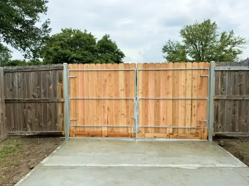 Wooden gate and fence, new light wood color, set in concrete, against gray sky.