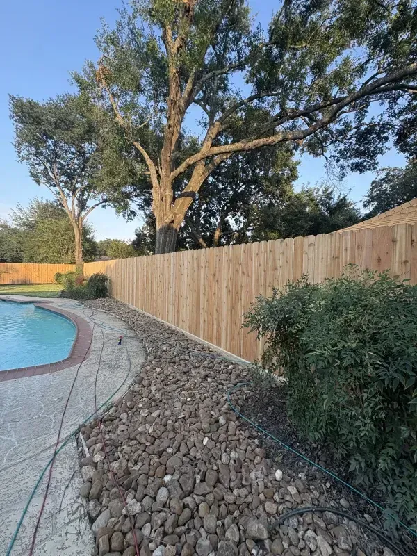 Wooden fence alongside a pool, with river rocks and greenery. Large tree in background.