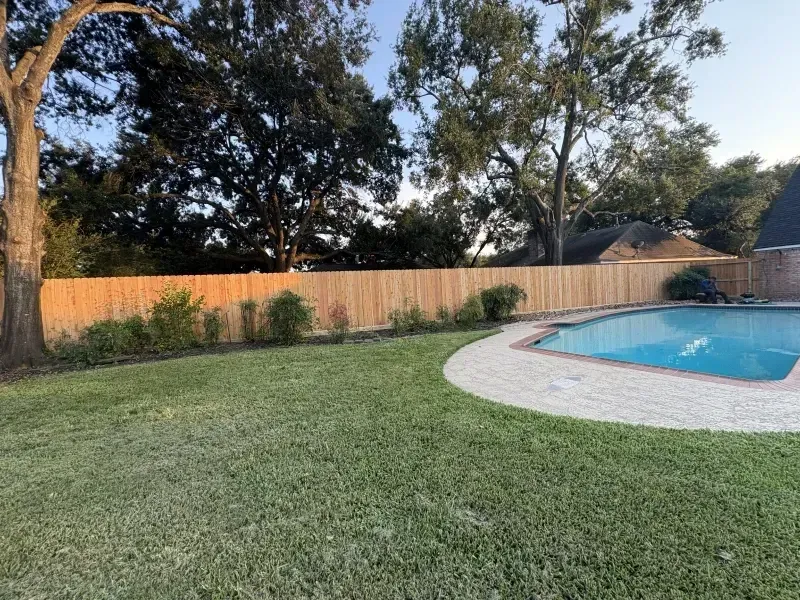 Backyard with a wooden fence, green lawn, pool, and trees.