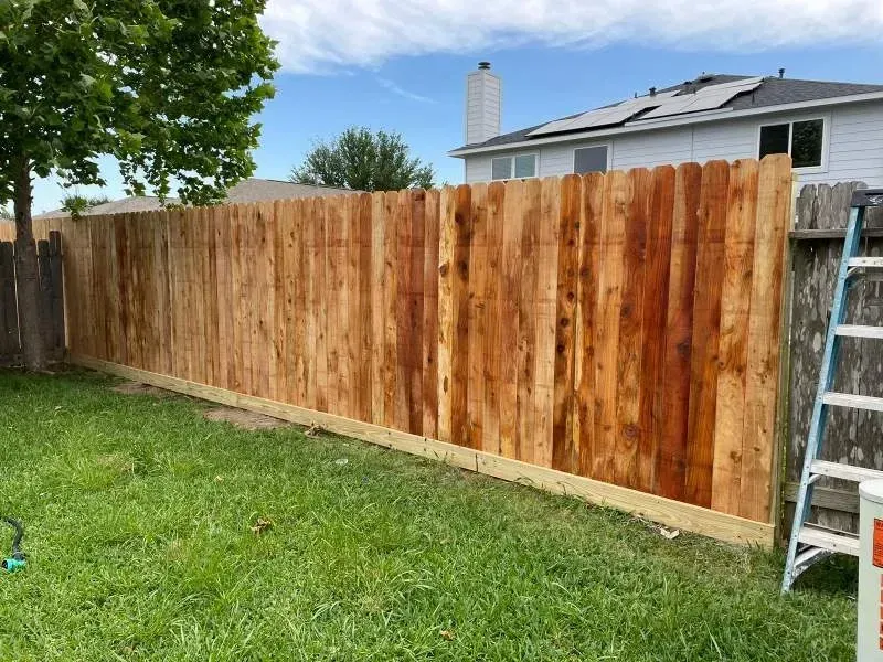 Wooden fence in a backyard with green grass, a house in the background, and a ladder.