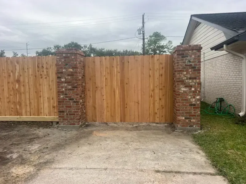 Wooden fence with brick columns forming a gate. A driveway leads up to it.