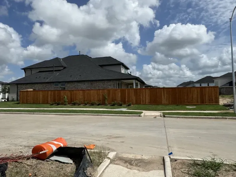 A new, wooden privacy fence surrounds a two-story house on a street under a cloudy sky.