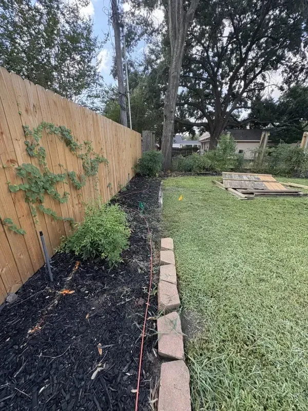 Backyard with a wooden fence, flowerbed with mulch, and grassy area with a brick border.