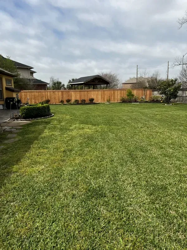 Green lawn with a wooden fence and a gazebo in the background on a cloudy day.