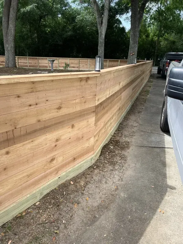 A long wooden fence, light brown color, borders a driveway next to a yard with trees.