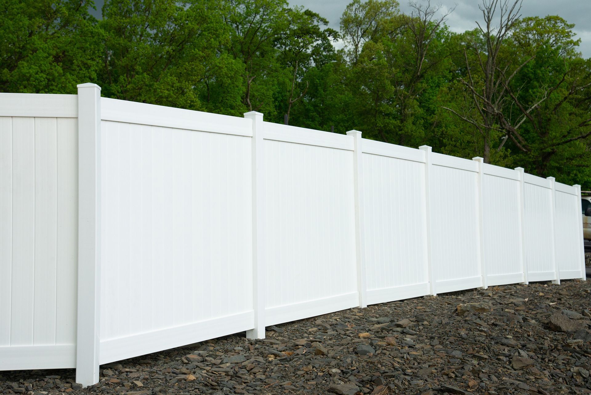 White vinyl fence along a rocky area with green trees in the background.