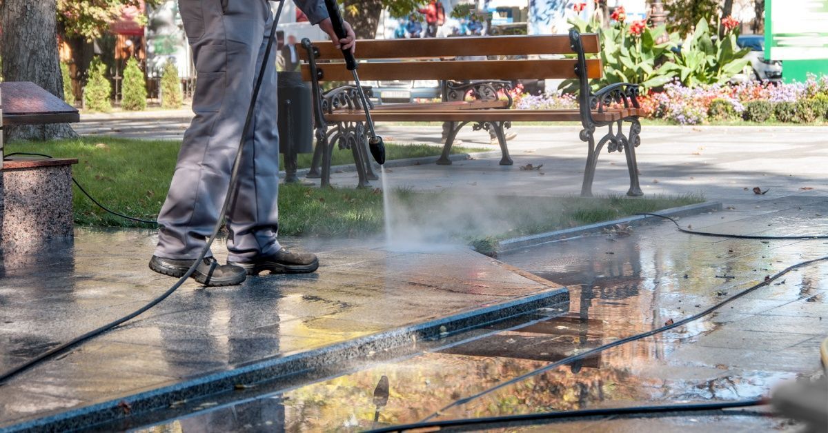 A person uses a pressure washer to clean pavement near a bench in an urban area. Water accumulates by the sidewalk.
