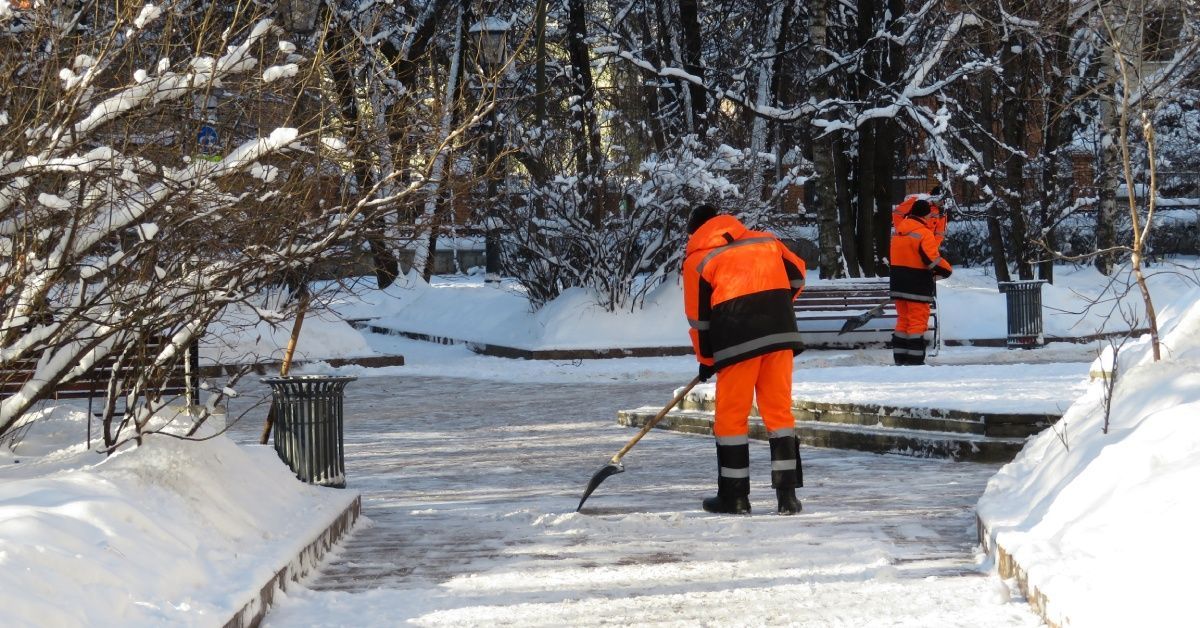 Two communal service workers clear snow and ice from the walkways at a public park with plastic shovels.