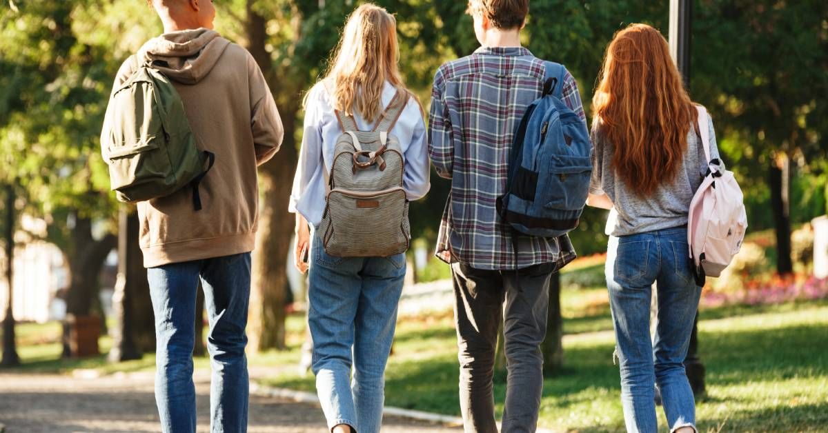 Four young students wearing backpacks walk down a paved path between trees, providing shade on a sun