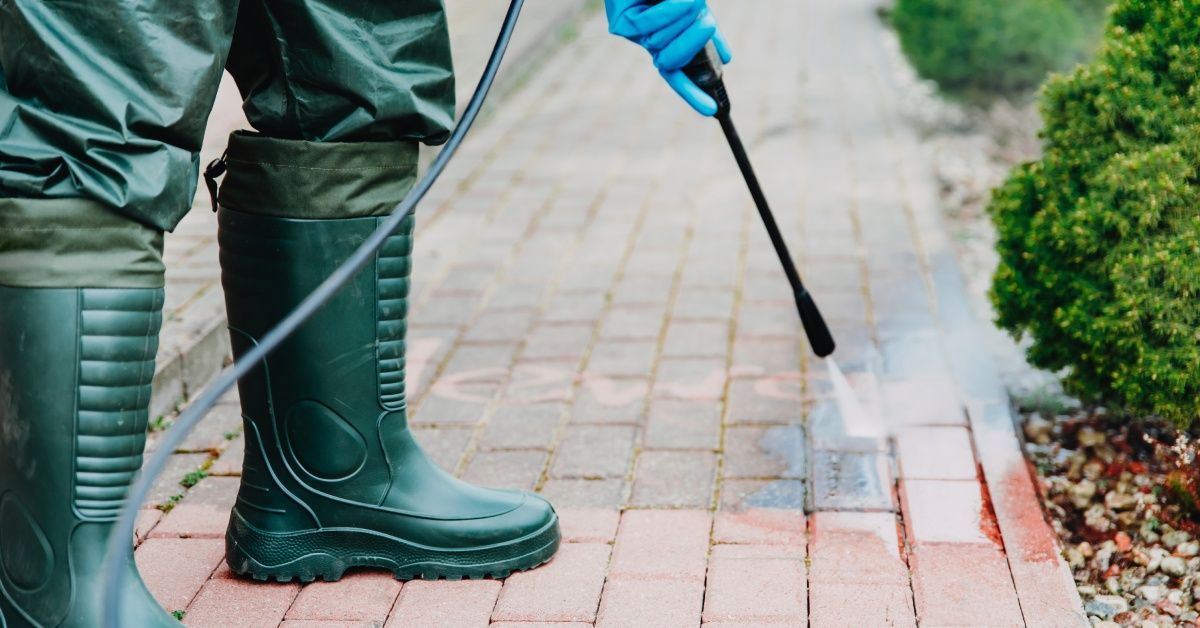 A maintenance worker uses a pressure washer gun to clean the stone paving crevices near small bushes.