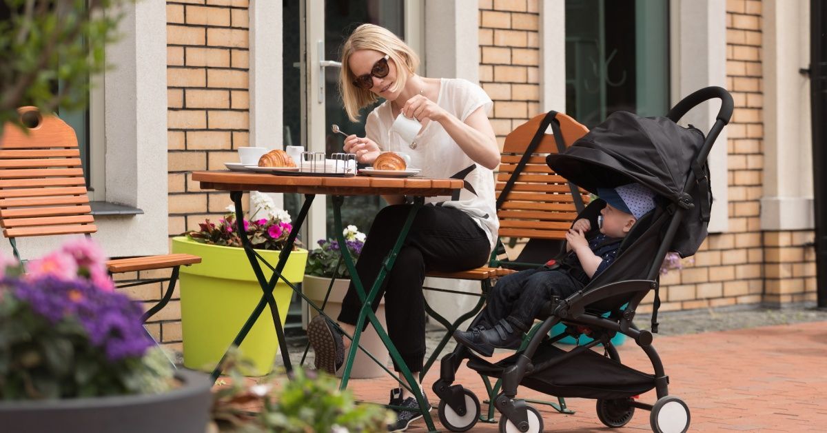 A mother wearing sunglasses sits at an outdoor cafe table with her infant in a stroller next to her.