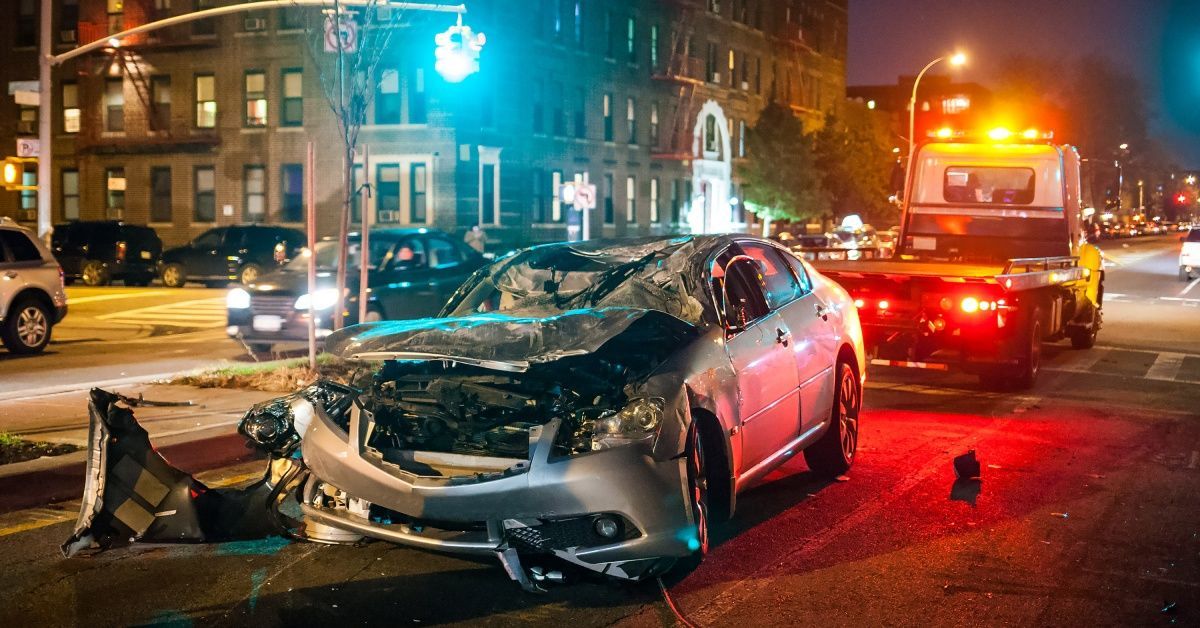 A crashed car sits at an intersection where a tow truck prepares to remove it. Pieces of debris are scattered around.