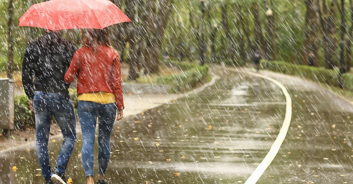 A couple walks along a tree-lined pathway during a heavy storm, with pouring rain. They share a red umbrella.