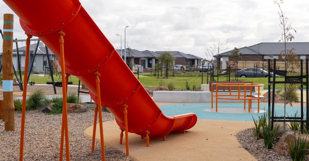 A playground with permeable paving has a large red slide in the foreground. Houses are in the background.