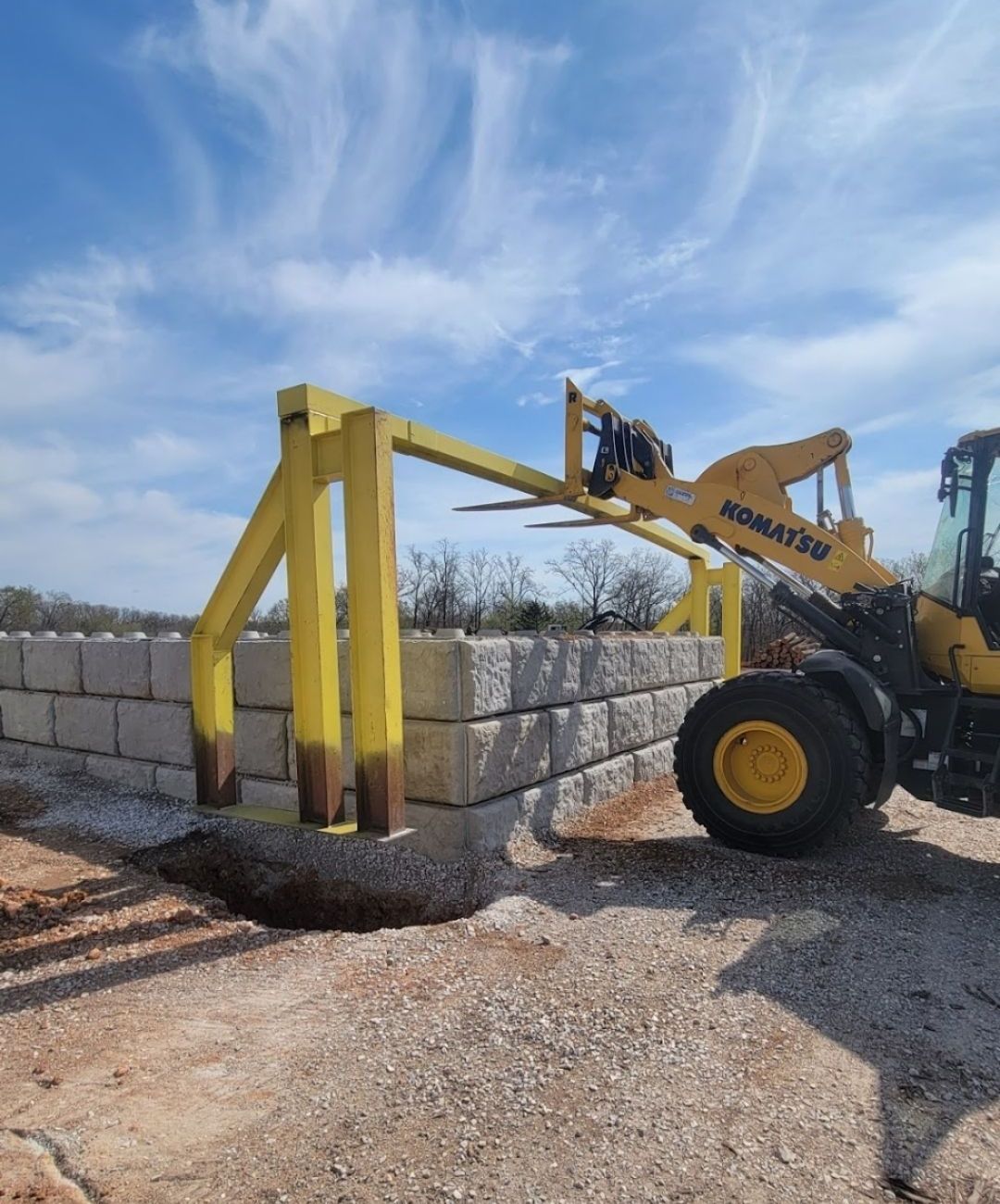 Yellow Komatsu forklift lifting a stack of gray concrete blocks with a specialized attachment. Sunny day.