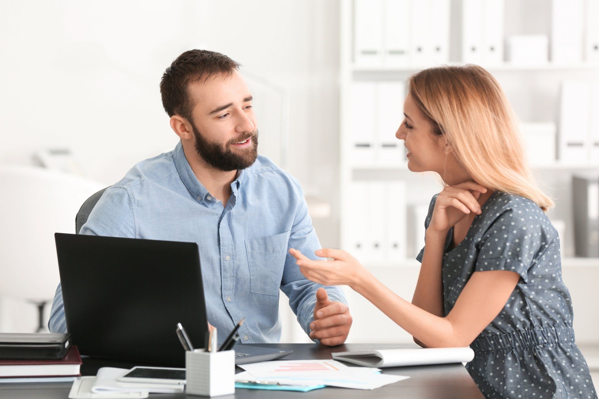 Two people talking at a desk with a laptop and papers in a bright office