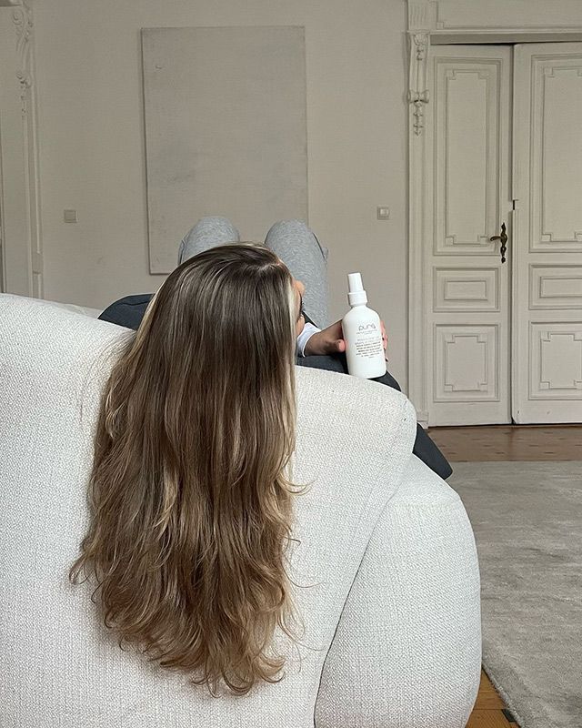 A Woman With Curly Blonde Hair Is Sitting On A Couch Holding A Spray Bottle — Sophisticut Hair Salon in Mysterton, QLD