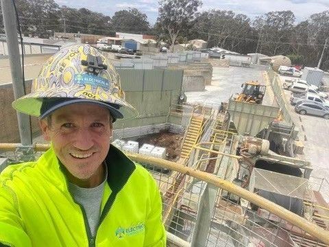 A Man Wearing a Hard Hat and A Yellow Jacket Is Standing in Front of A Construction Site — Bay Coast Electrical & Control Solutions in Maloneys Beach, NSW