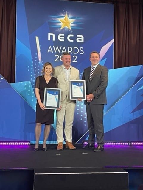 Three People Stand On A Stage In Front Of A Neca Awards Sign — BayCoast Electrical & Control Solutions in Maloneys Beach, NSW