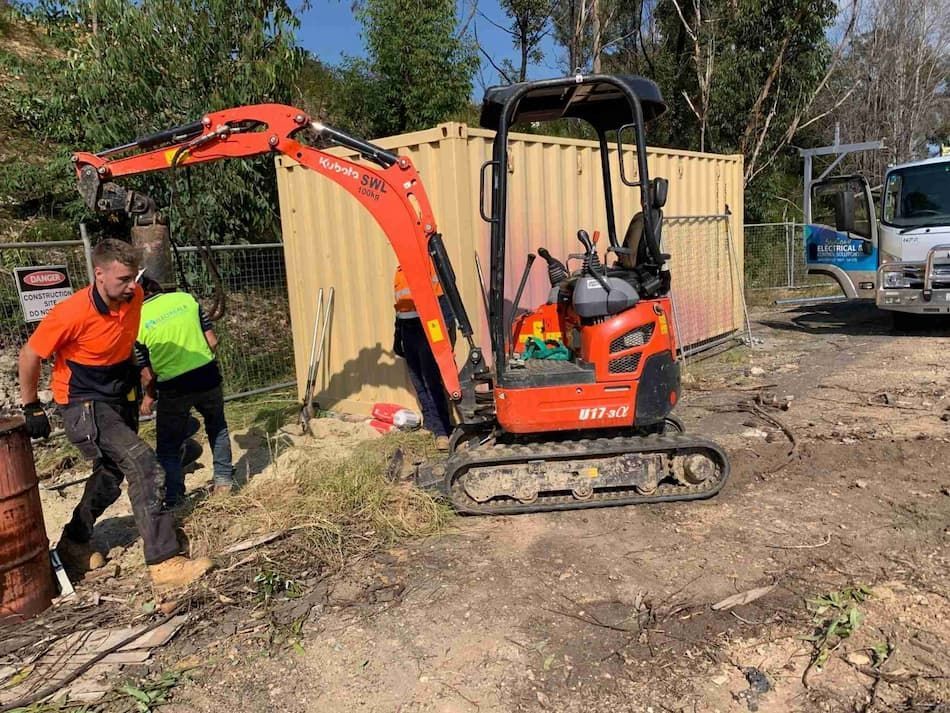 A Man Is Standing Next to A Small Excavator in A Dirt Field — Bay Coast Electrical & Control Solutions in Maloneys Beach, NSW