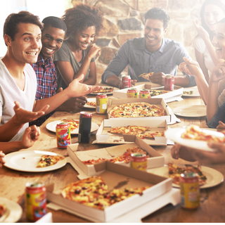 Group of friends smiling and eating pizza at a table, with pizza boxes, soda cans, and plates.