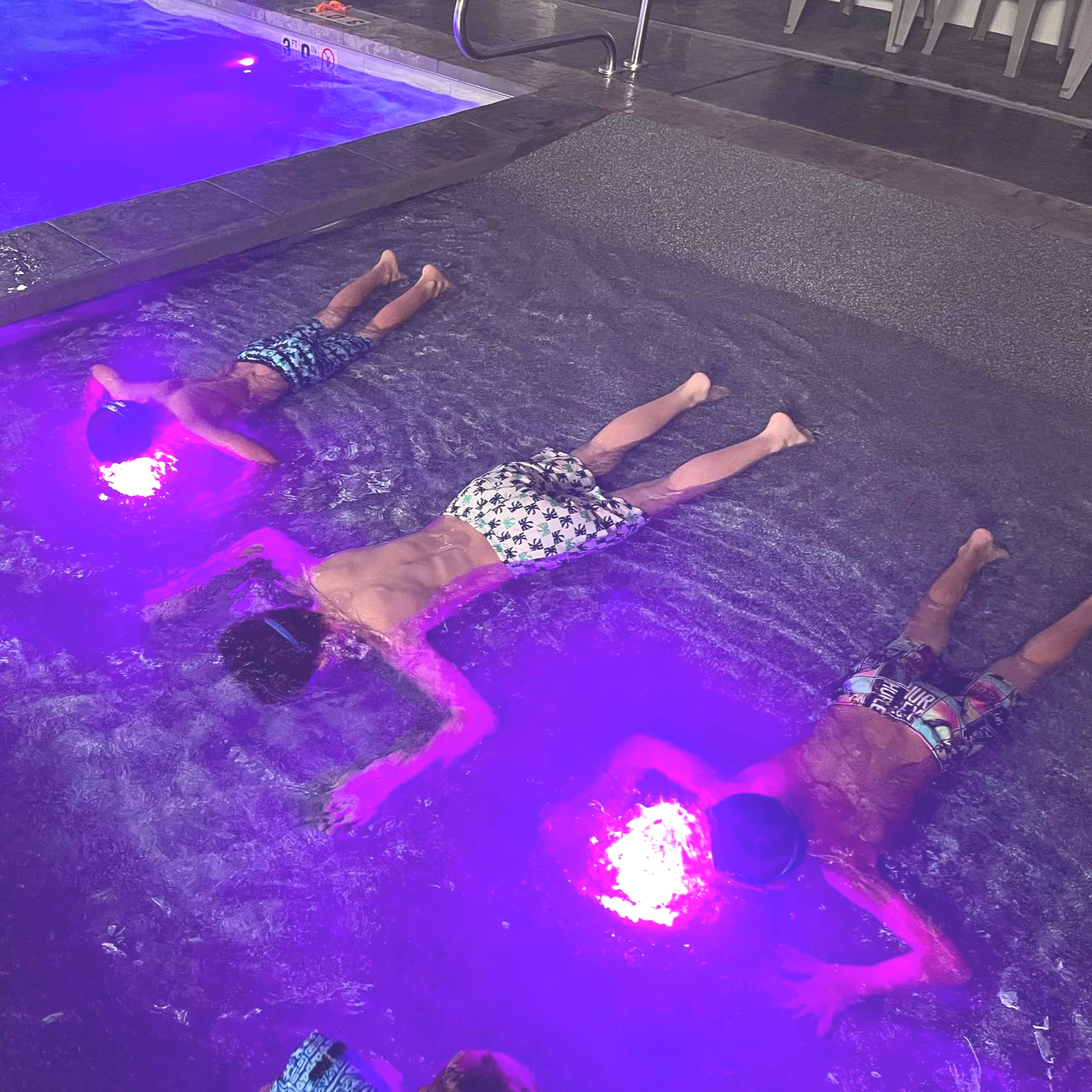 Three boys floating in a pool with purple light. They are wearing swim trunks and appear relaxed.