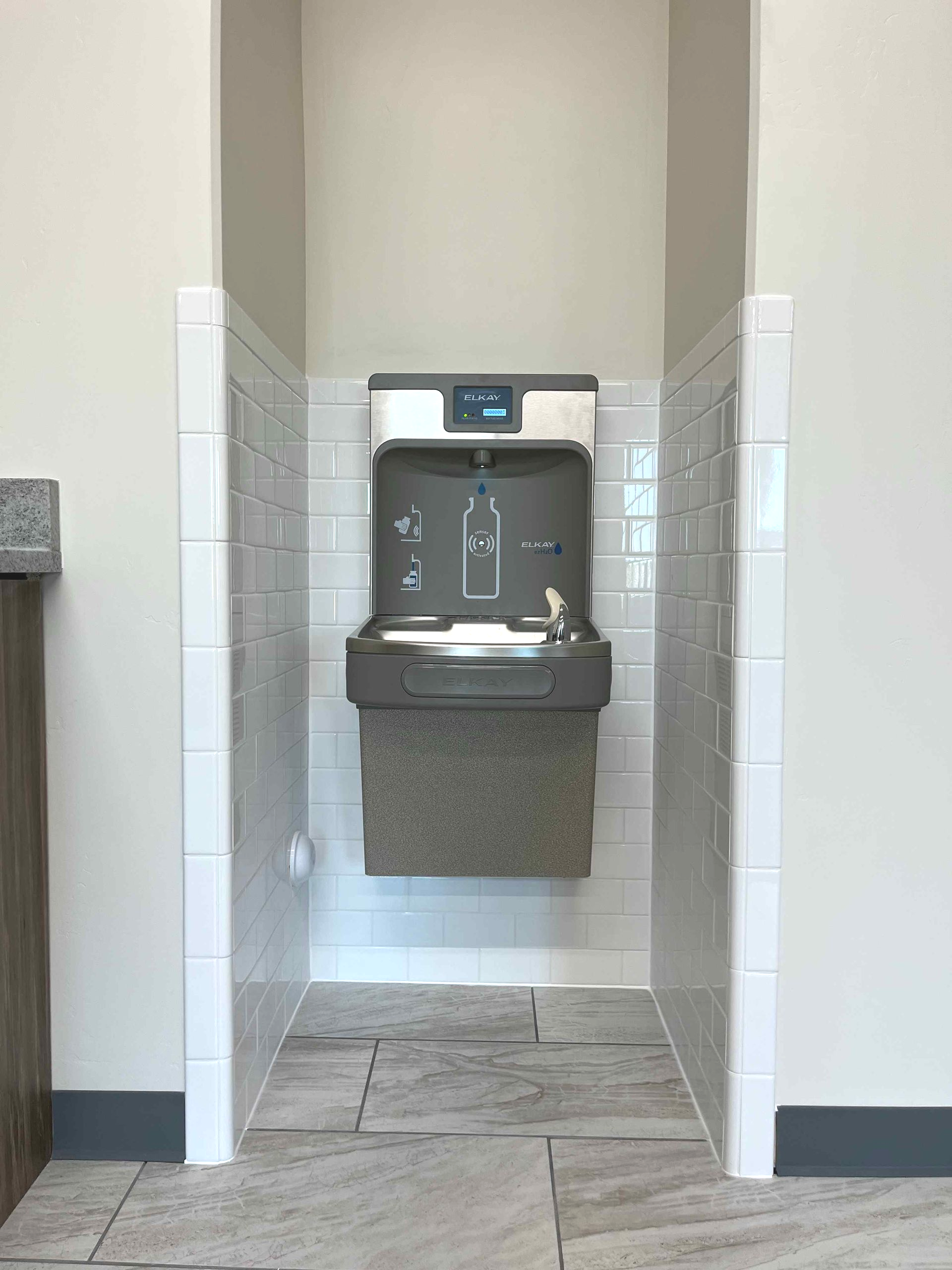 Water fountain in a tiled alcove. Gray and white, bottle filler, accessible design.