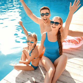 A happy family of three by a pool. A man and woman raise their arms with excitement, while a child sits smiling, all wearing sunglasses.