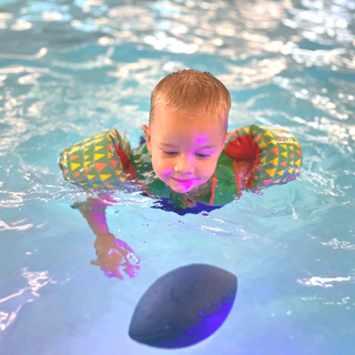 Boy in a pool wearing arm floaties, looking at a submerged football.