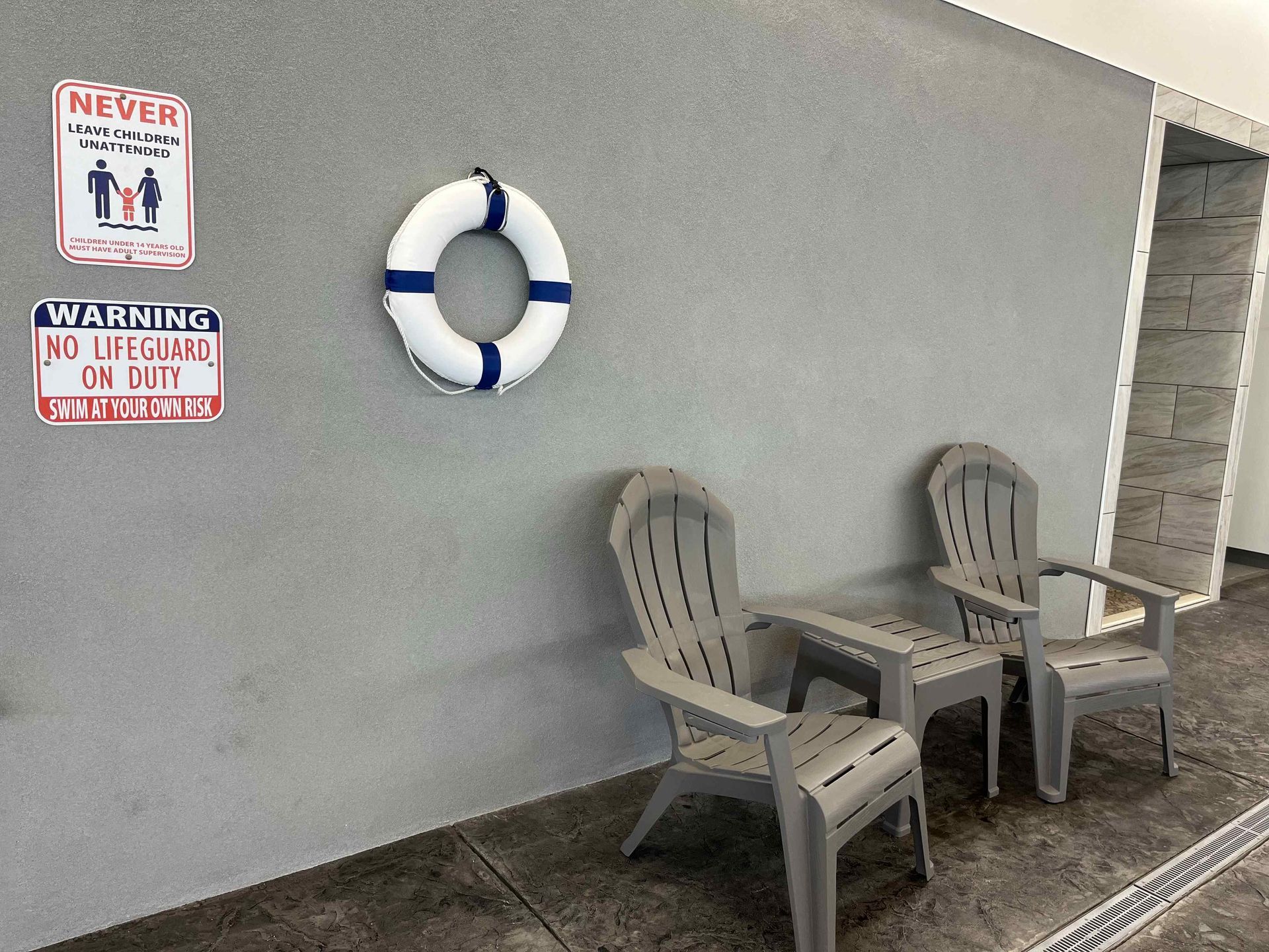 Two gray chairs, a life ring, and safety signs hang on a textured gray wall next to a pool.