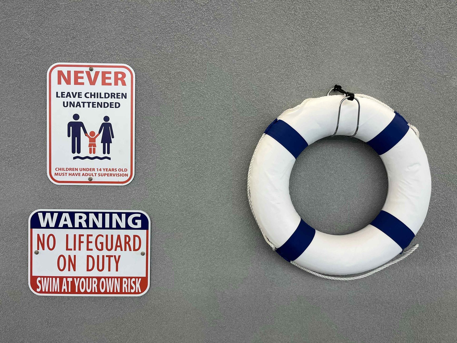 Signs and a life preserver on a grey wall: warning, no lifeguard on duty; never leave children unattended.
