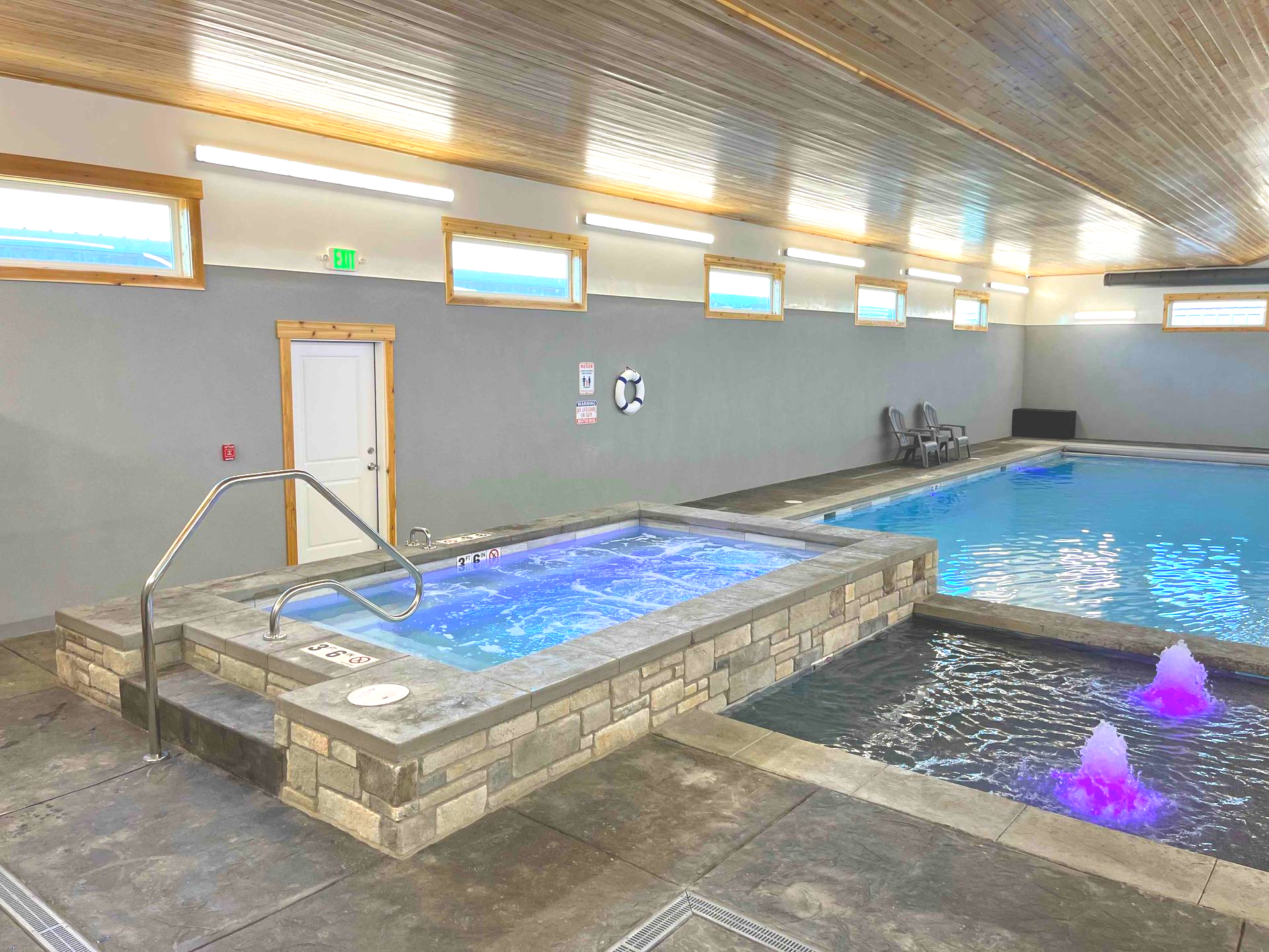 Indoor pool with a hot tub, fountains, and windows. Gray walls and light blue water.