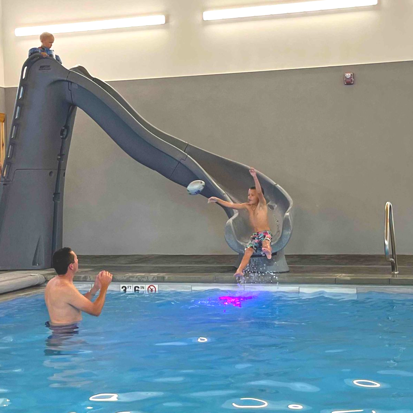 Boy slides into a pool, splashing as a man in the water reaches. Another child is at the top of the slide.