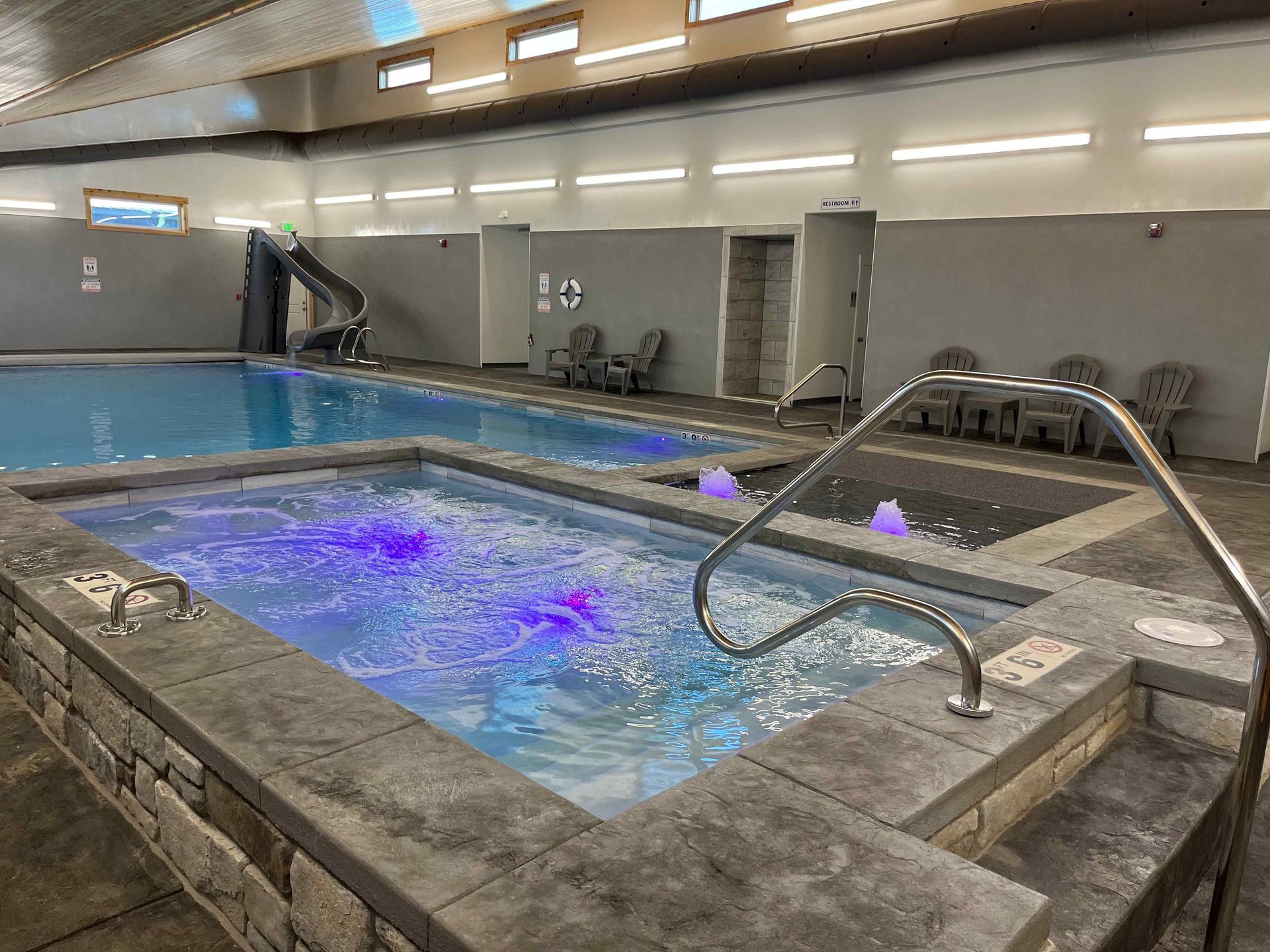 Indoor pool and hot tub with blue lights, seating, and a slide.