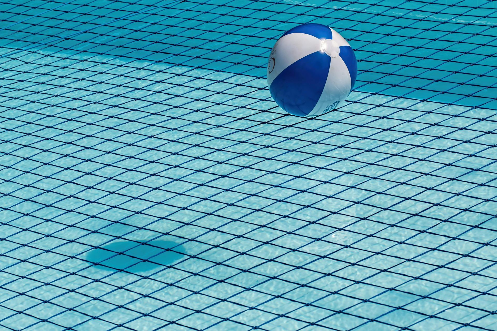 A blue and white beach ball floats in a sunlit swimming pool, casting a shadow on the tiled floor.