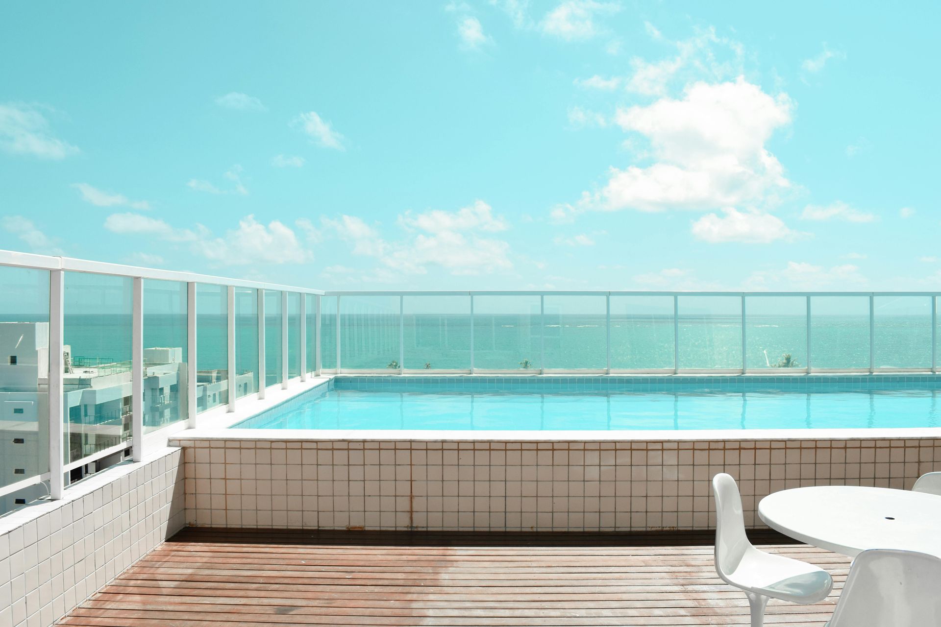Rooftop pool with ocean view under a bright blue sky.  White tables and chairs on wooden deck.