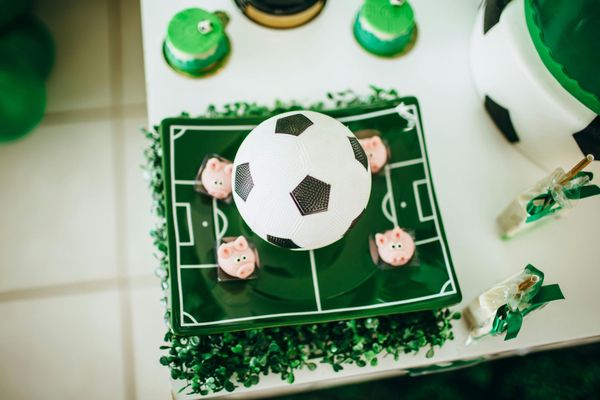 Soccer-themed cake with a soccer ball on top, decorated with a green field and small pig-shaped treats.