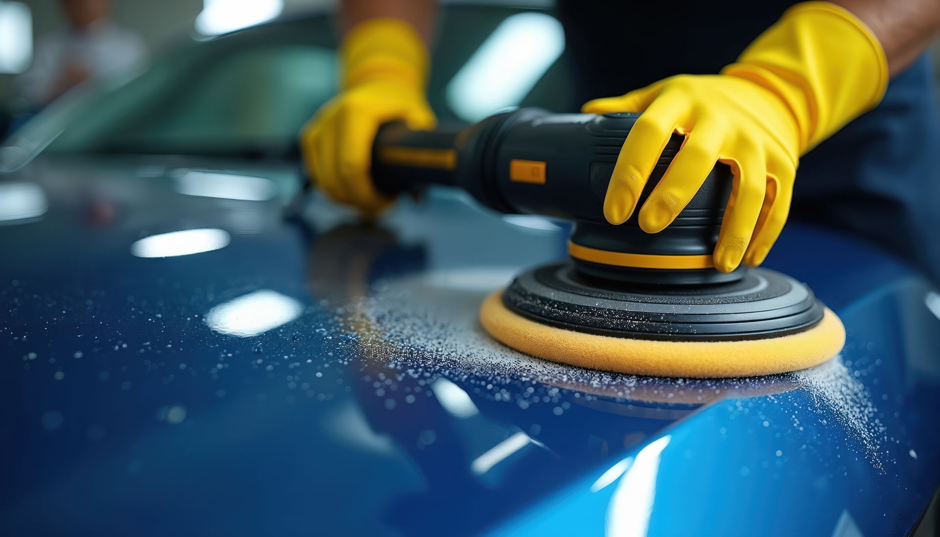 Person in yellow gloves polishing a blue car with an electric buffer.