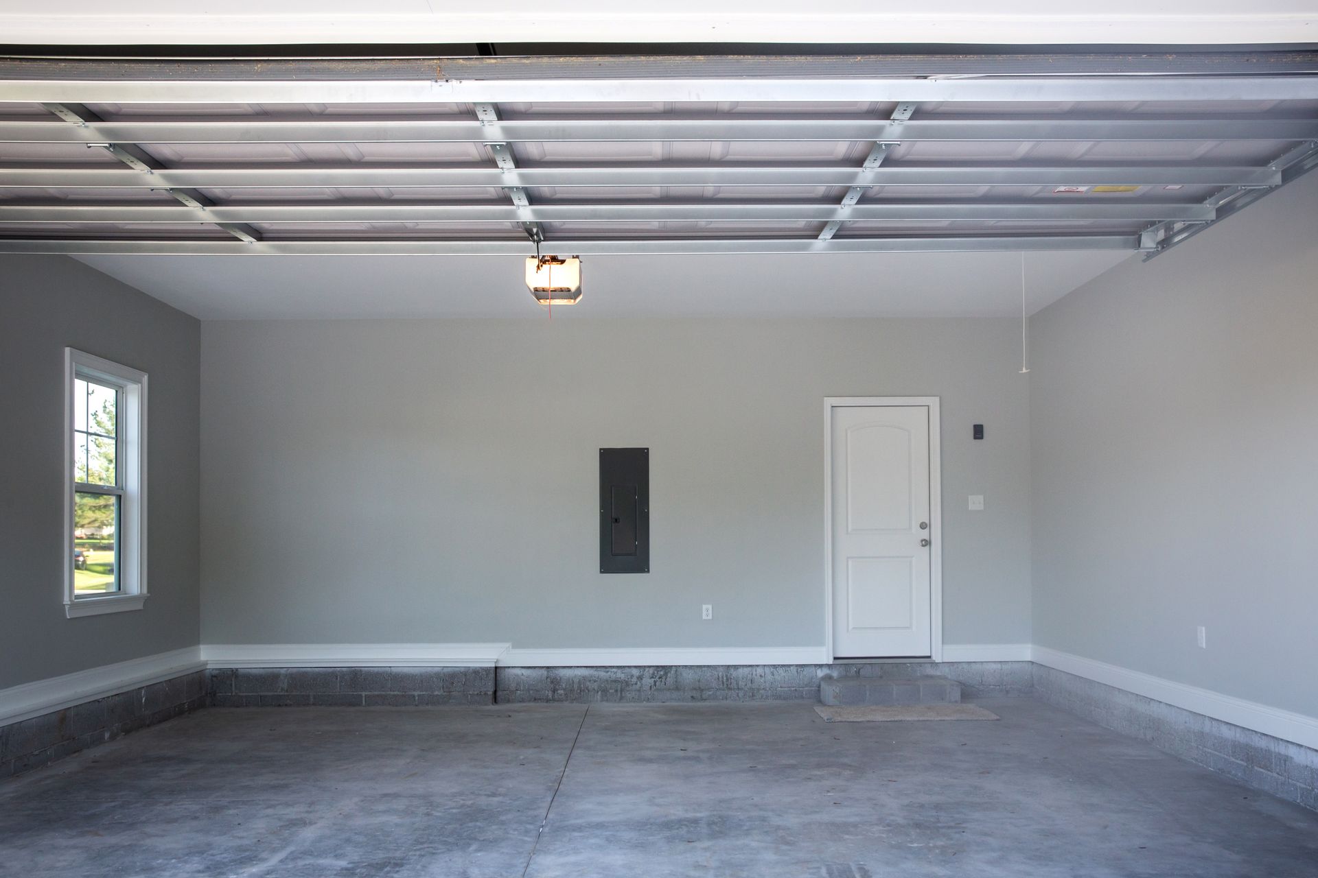 Empty gray garage with concrete floor, window, white door, and ceiling light.