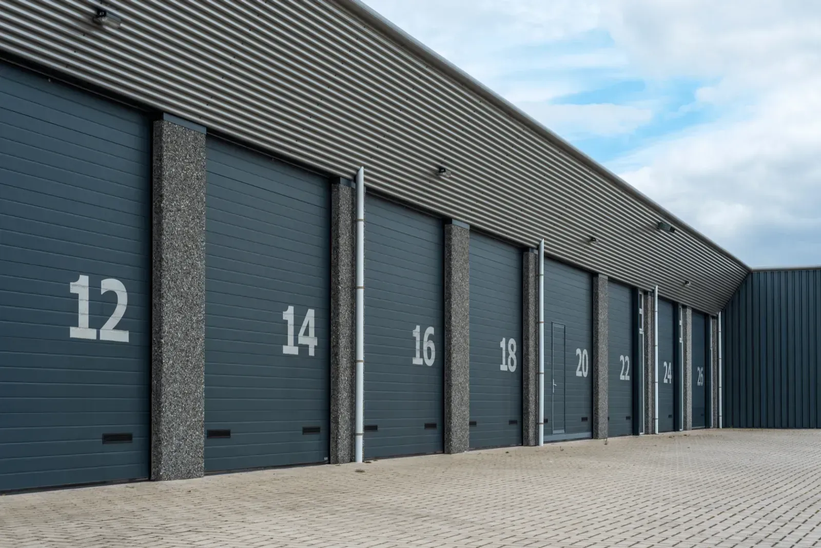 Row of gray industrial warehouse doors with white numbers on a paved lot under a cloudy sky
