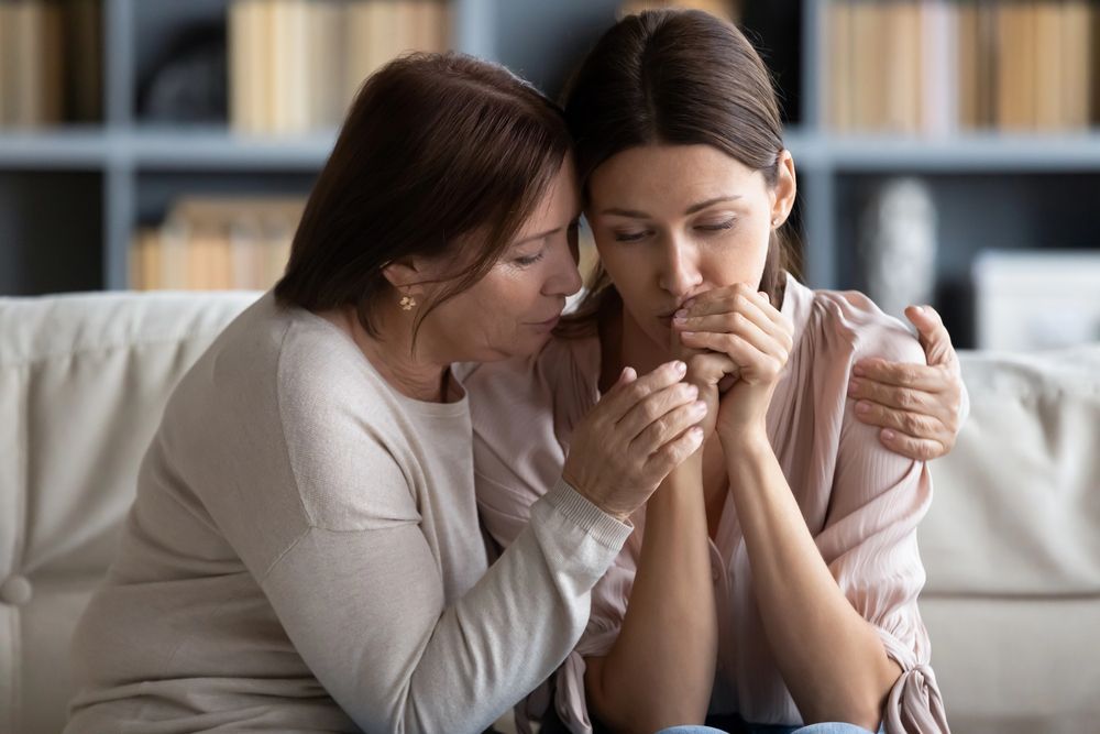 Woman comforts another, both seated, holding hands, with concerned expressions.