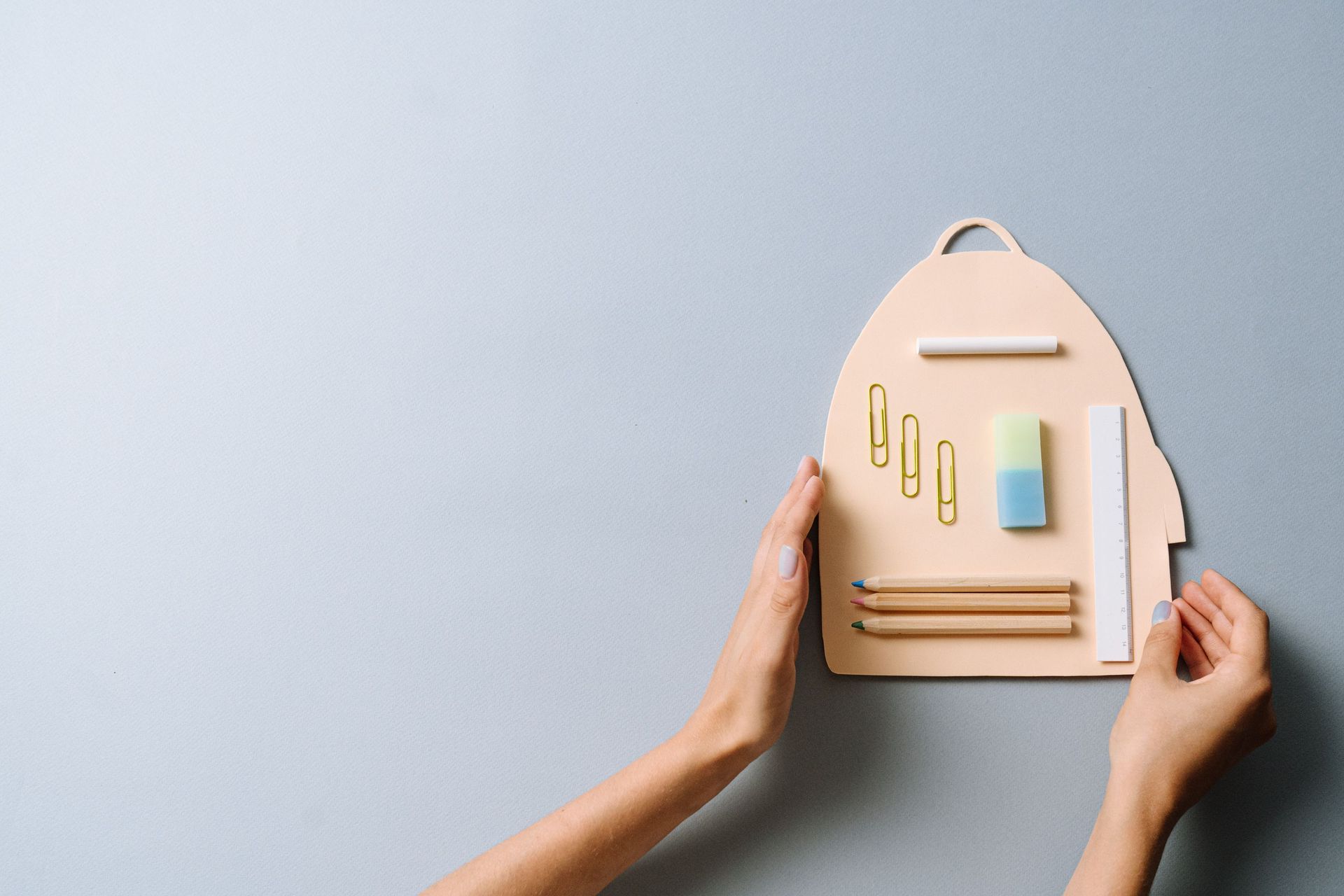Hands holding a small beige backpack with stationery on a light gray background