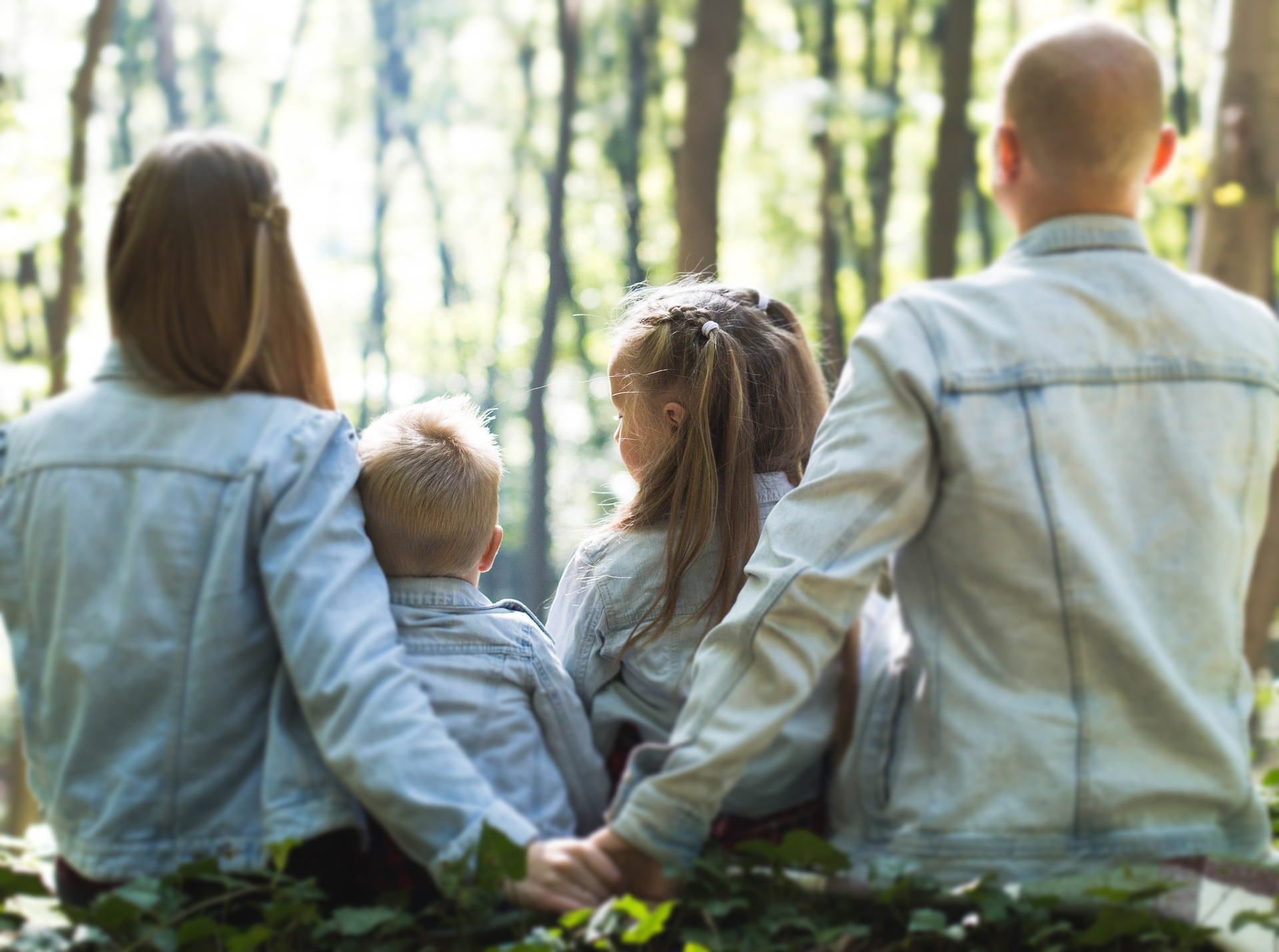 Family in matching denim jackets, backs to the camera, looking into a green forest.
